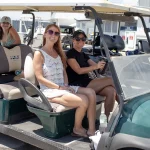 Photo of adults behind the wheel of a gold cart and two children on the rear most seat without seatbelts on.