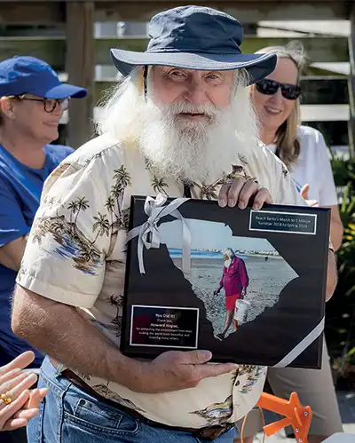 Howard Hogue holding a plaque reading at top, right, "Beach Santa's March to 1 Million. Summer 2018 to Spring 2024."