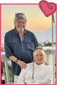 Older couple smiling in front of camera with a picturesque sunset in the background.